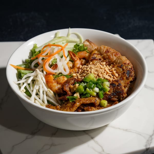 A white bowl of Vietnamese vermicelli noodles with grilled pork, shrimp, and fresh vegetables.