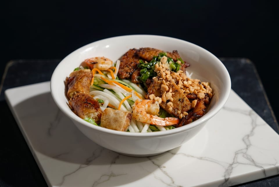 A white bowl of Vietnamese vermicelli noodles with grilled pork, shrimp, egg rolls, and peanuts.